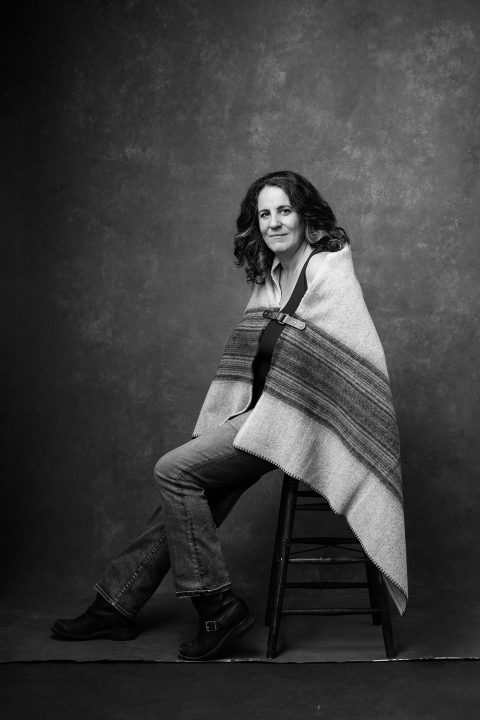 A black and white portrait of a woman wrapped in a blanket, leaning on a stool