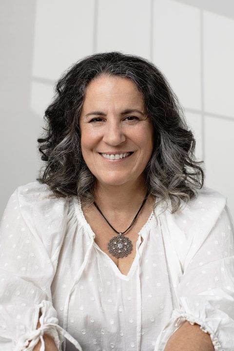 A woman in her 50s, smiling, wearing white, with a white background and window shadows