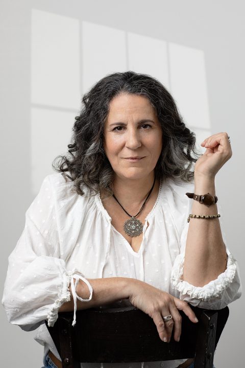 A woman in her 50s, wearing white with a white background and window shadows behind her