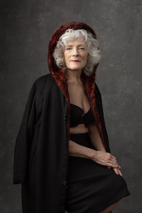 Portrait of Jill for Unforgettable: the Over 50 Revolution.  She has white hair over 70, wearing a black bra and skirt, draped in a black hooded coat.