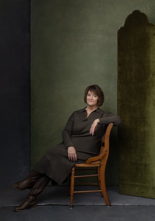 Susan, seated wearing olive dress in front of green backdrop and velvet room divider