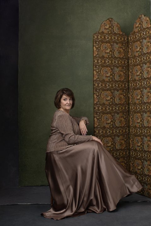 Susan, seated, wearing a copper-colored satin gown in front of a green, hand-painted backdrop, next to an antique room divider
