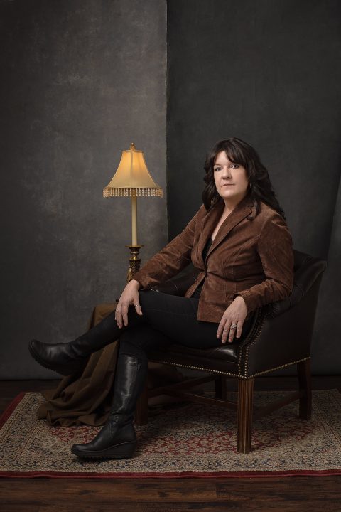 Portrait of Kree for Unforgettable: the Over 50 Revolution. She is sitting in a leather chair, with vintage rug and lamp