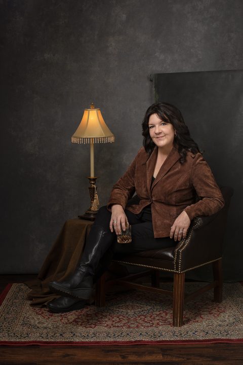 Portrait of Kree for Unforgettable: the Over 50 Revolution.  She is sitting in a leather chair, holding a glass of whiskey