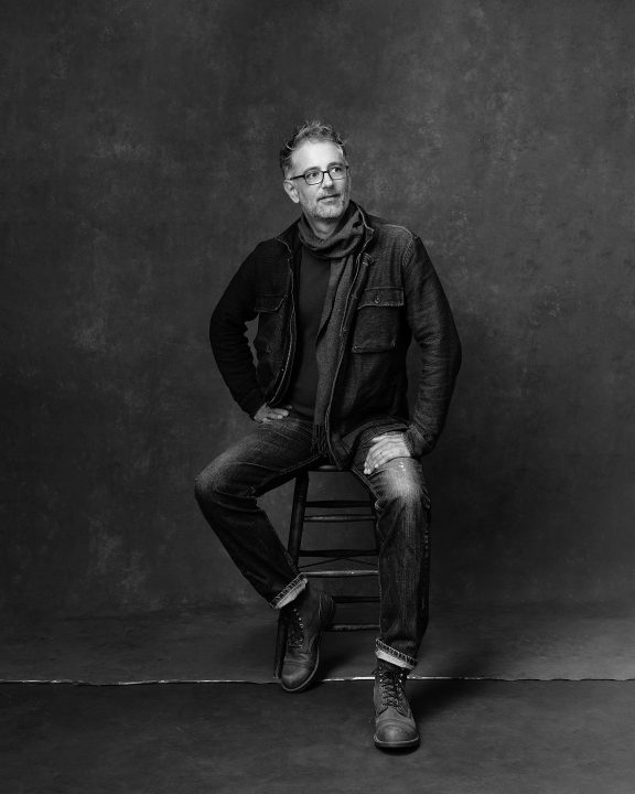 Casual, black and white portrait of man sitting on stool, wearing jeans and scarf