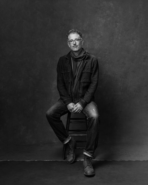 Black and white portrait of a man sitting on a stool, wearing jeans and a scarf