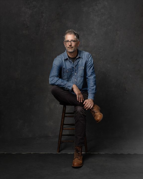 Casual studio portrait of man sitting on stool, wearing denim shirt