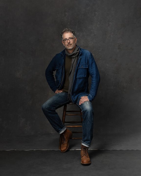 Casual portrait of a man sitting on a stool, wearing jeans and a scarf