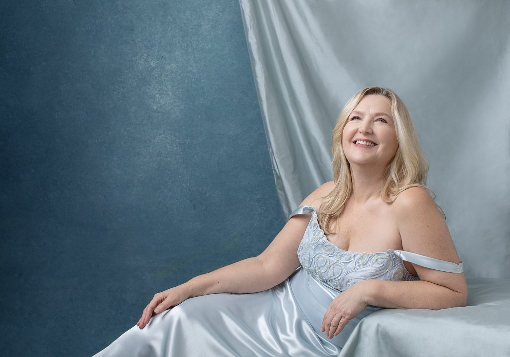 Portrait of a woman over 50, reclining and laughing, wearing a light blue gown, in front of a blue layered backdrop