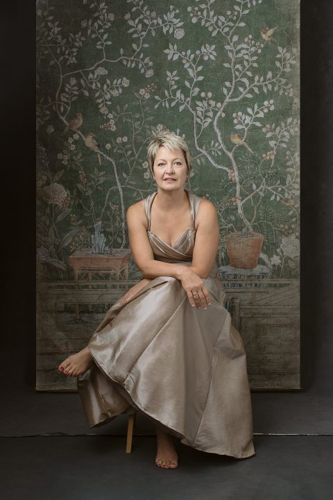 Portrait of Carol sitting in front of an antique garden-inspired tapestry, wearing a champagne silk dress for Unforgettable: the Over 50 Revolution at Maundy Mitchell Photography