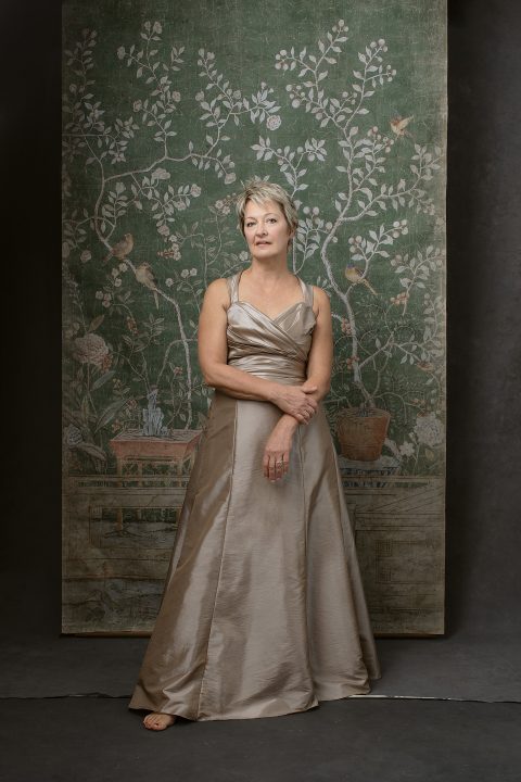 Carol, standing in front of antique floral tapestry wearing a champagne silk dress for her portrait session for Unforgettable: the Over 50 Revolution 