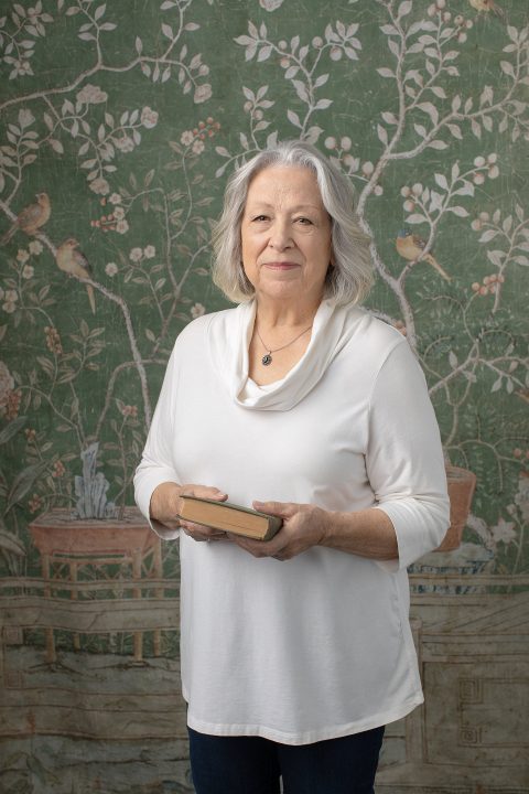 Photo of Liz from her Over 50 Revolution portrait experience, holding a book in front of a vintage tapestry