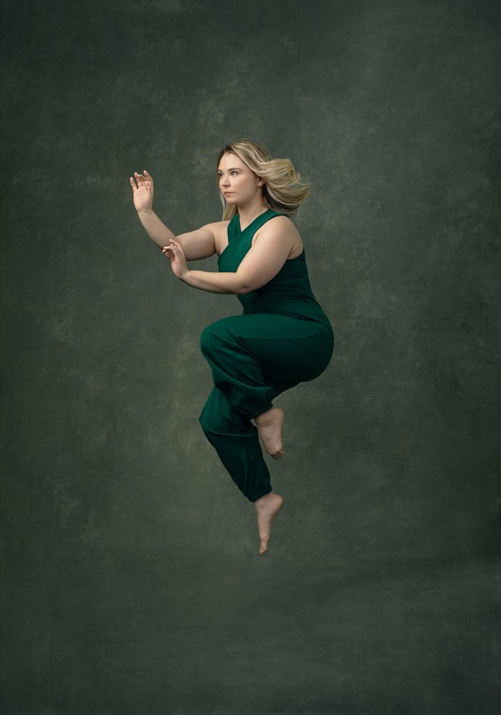 Little, Fierce - dancer Suzanna in midair, wearing a green jumpsuit