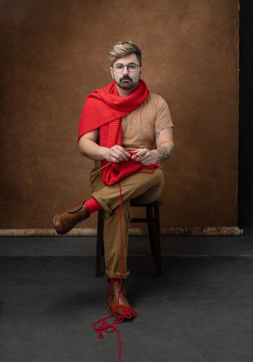 "Knitted Together"- Michael - A portrait of a man seated, knitting a red scarf that is wrapped around his neck as the yarn enters the foreground and comes out of the frame. ©Maundy Mitchell 2023