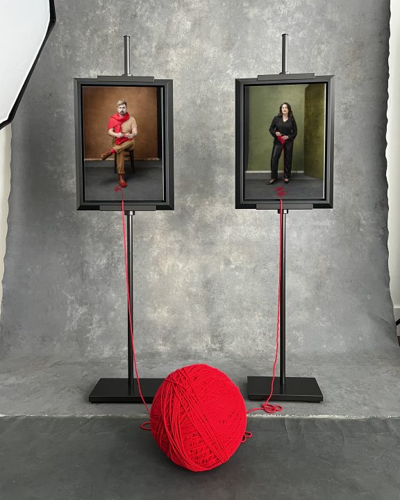 A cell phone photo of the Knitted Together project beginning, with two portraits printed on canvas, framed, on easels, with strands of yarn connecting them to a large ball.