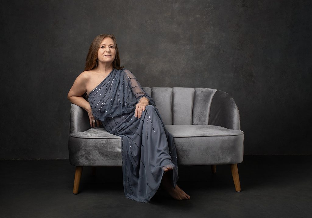 A portrait of Kathy G. for Extraordinary: the Over 50 Revolution. She is wearing a beaded gown, sitting on a velvet sofa, and barefoot.