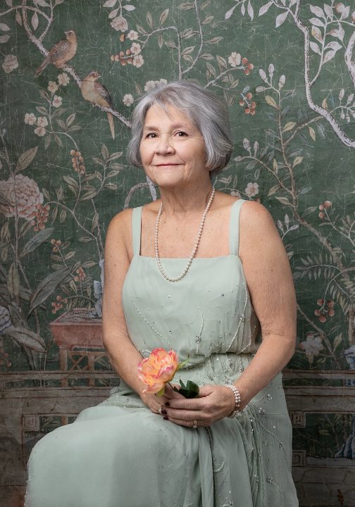 Portrait from Extraordinary: the Over 50 Revolution - Rebecca seated, wearing a pale green dress in front of an antique green floral tapestry