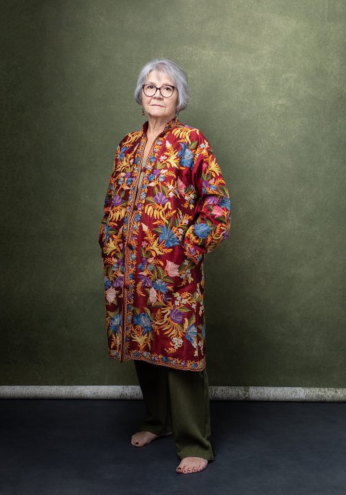 Portrait from Extraordinary: the Over 50 Revolution - Rebecca wearing a colorful embroidered coat in front of a green hand-painted backdrop