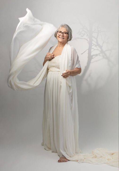 Portrait from Extraordinary: the Over 50 Revolution - Rebecca smiling, wearing a white outfit with flowing fabric, with shadows of trees in the background