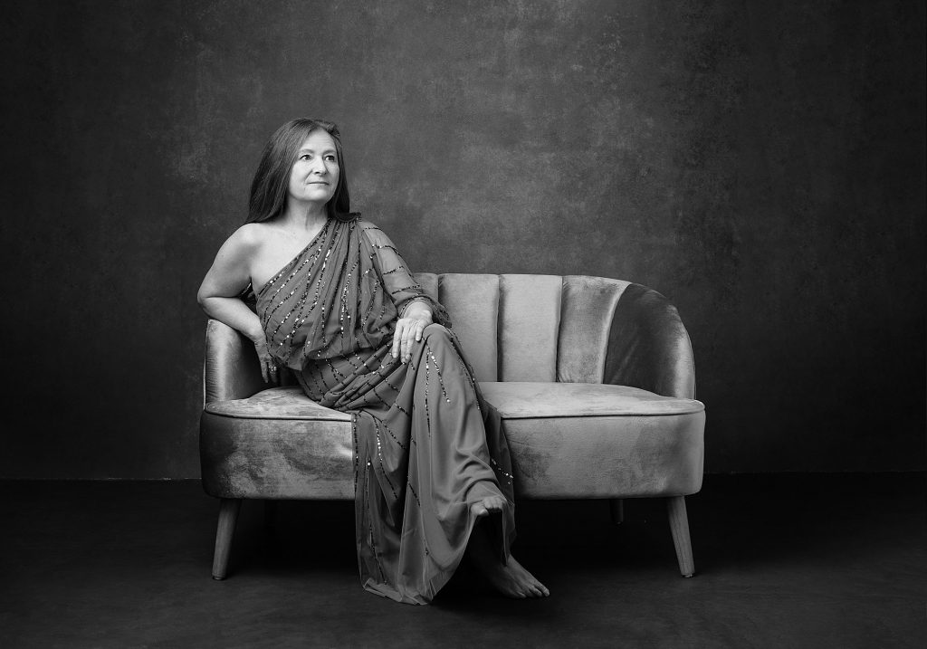 A black and white portrait of Kathy G. wearing a beaded gown and sitting on a velvet sofa, looking away. Part of her portrait experience for the Over 50 Revolution at Maundy Mitchell Photography