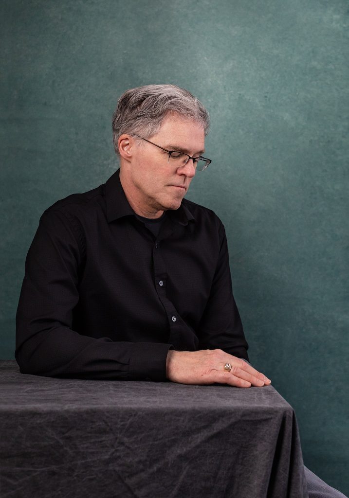 Photo of Boston area portrait painter Paul Mock looking down, sitting at table in front of teal background