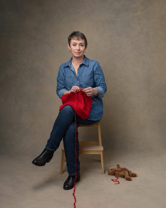 Knitted Together: Shirley
A portrait of a woman knitting with red yarn and a bear on the floor beside her