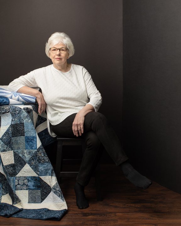 NH Quilter Susan Hofsetter sitting with a stack of quilts in front of a black background