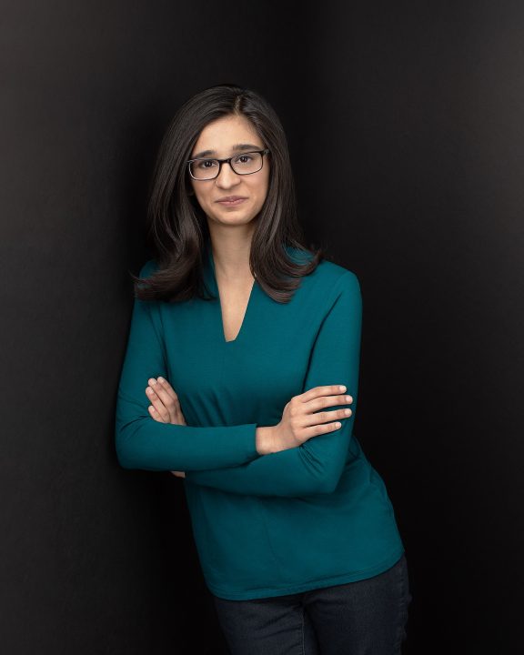 Riyah Patel leaning arms crossed in front of black background