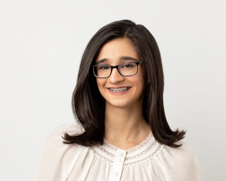 A headshot of Riyah Patel. She is wearing white in front of a white background. She is smiling, showing braces
