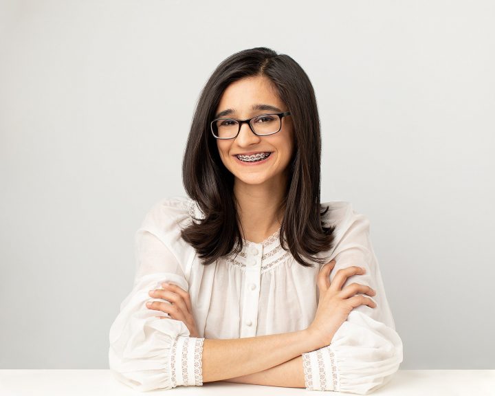 Riyah Patel seated at table smiling - wearing white top in front of white background