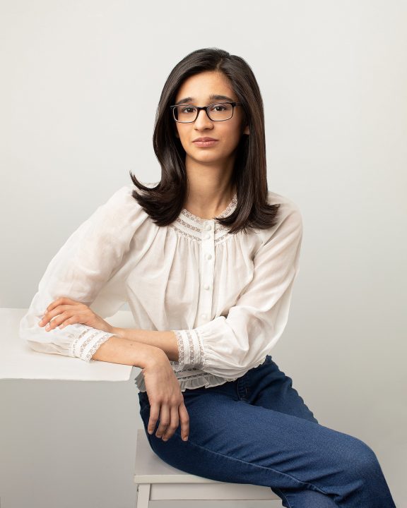 Riyah Patel seated beside a table - wearing white top in front of white background