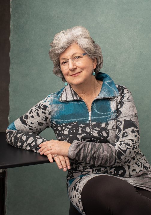 Over 50 Revolution portrait of Susan C. wearing patterned tunic in front of teal background. She is seated and leaning on a table