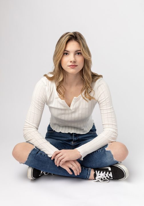 Senior photo of Brianna, sitting on floor, wearing jeans and sneakers, in front of white background
