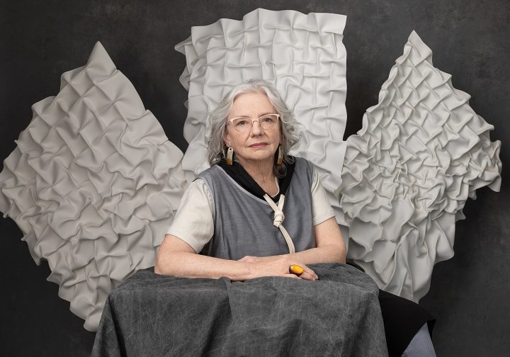 Portrait of Kathy, seated, with her artwork hanging behind her.