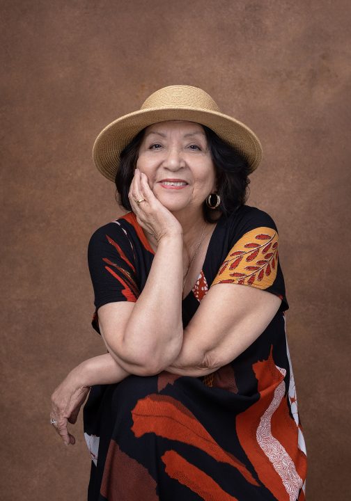 Maria, seated, wearing a hat, smiling for her portrait session for Extraordinary: the Over 50 Revolution