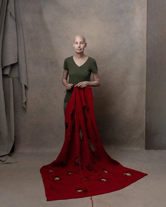 Portrait of Jody for Knitted Together, Part V. She is standing, holding knitted needles that are attached to a red knitted blanket. The blanket is riddled with holes. A single strand of red yarn leaves the frame from the bottom center.