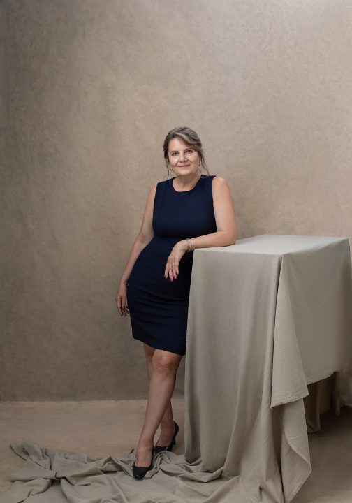 Portrait of Beth, standing, leaning on a table draped with beige fabric in front of a beige, hand-painted backdrop