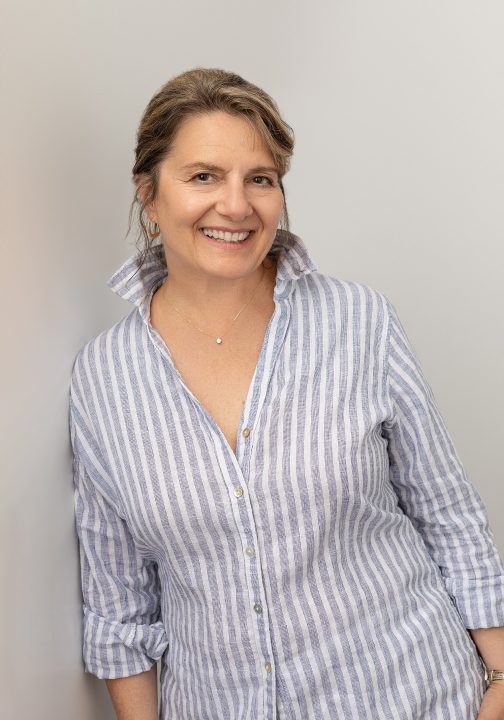 Casual portrait of Beth from her photo session for Extraordinary: the Over 50 Revolution. She is wearing a striped shirt, leaning against a light gray wall