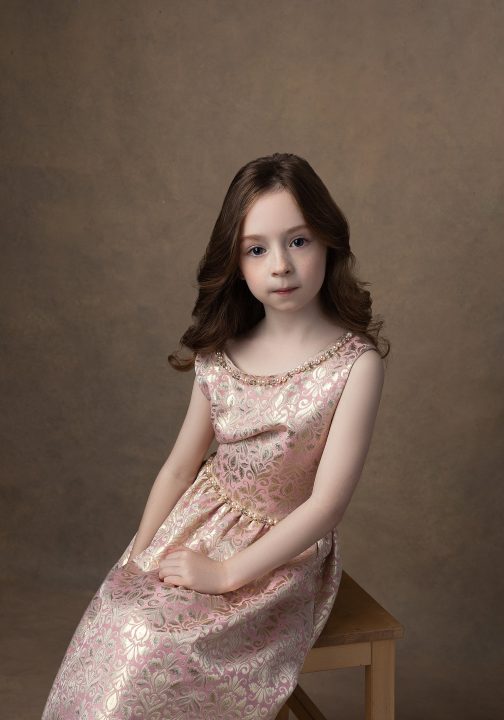 Portrait of a young girl seated, leaning towards the camera, wearing a gold and pink brocade dress.