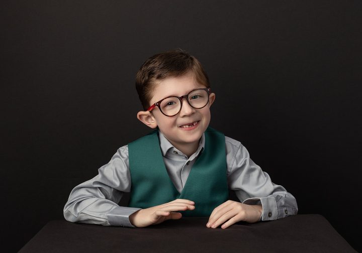 Photo of a young boy sitting at a table, wearing glasses and a green vest, smiling and drumming with his hands