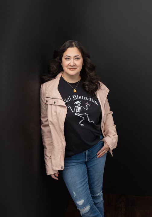 Portrait of a woman with dark hair, leaning and wearing a pink leather jacket and jeans, with a black background