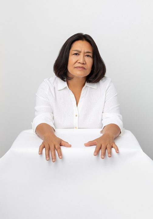 A portrait of Lisa wearing white with a white background, seated, with her hands reaching toward the camera