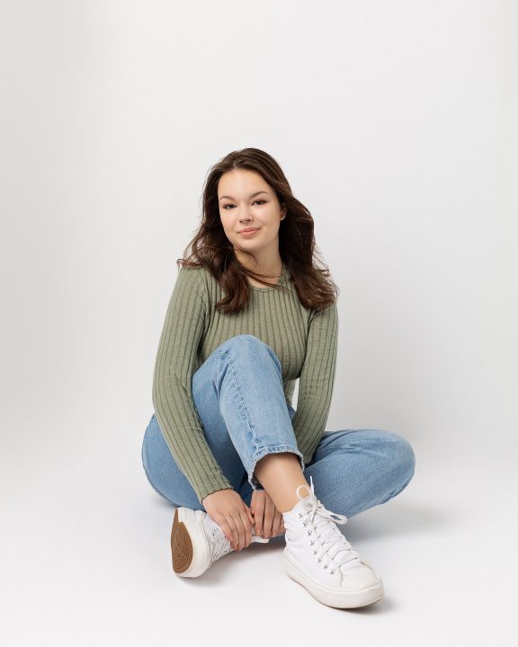 photo of a young actor sitting on the floor with a white background. She is wearing a sweater, jeans, and sneakers.