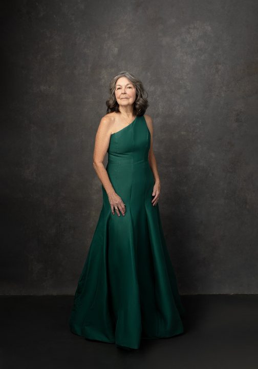 Portrait of Deb, standing, wearing a one-shoulder, green Halston gown