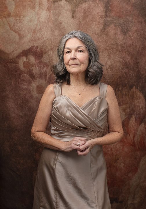 Portrait of Deb for the Over 50 Revolution. She is wearing a beige silk dress in front of a floral backdrop