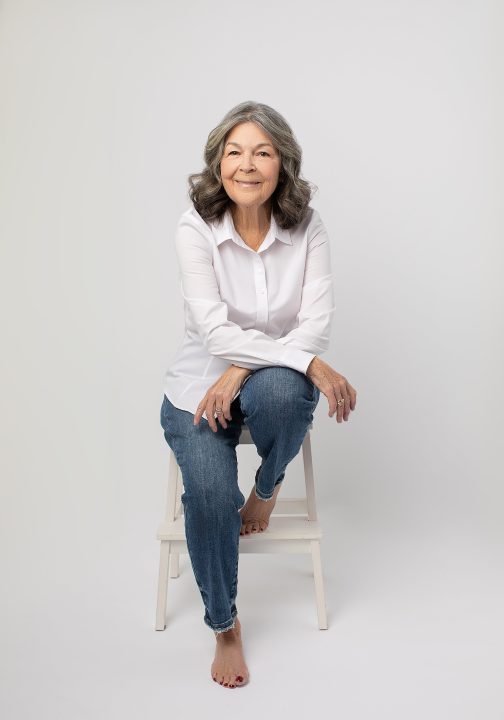 Photo of Deb from her portrait session for Extraordinary: the Over 50 Revolution. She is seated and barefoot, wearing a white shirt and jeans