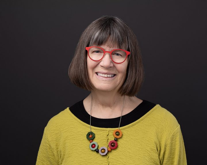New headshot for NH artist Marian Federspiel. She is wearing a colorful outfit with red glasses, in front of a black background.