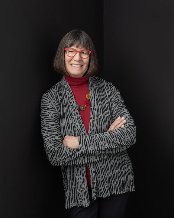 New headshot for NH artist Marian Federspiel. She is wearing a colorful outfit with red glasses, in front of a black background.