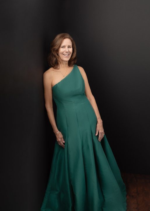 Robin wearing a one-shoulder green gown leaning in front of a black background for the Over 50 Revolution
