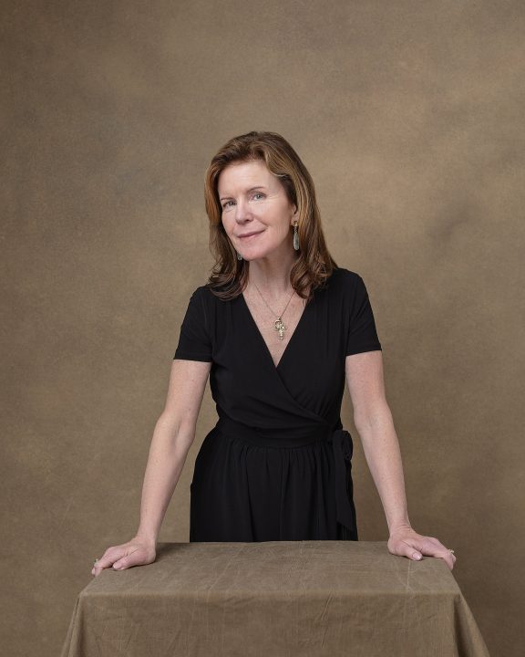 A portrait of Debra, wearing a black jumpsuit, leaning on a table - from her photo session for The Over 50 Revolution